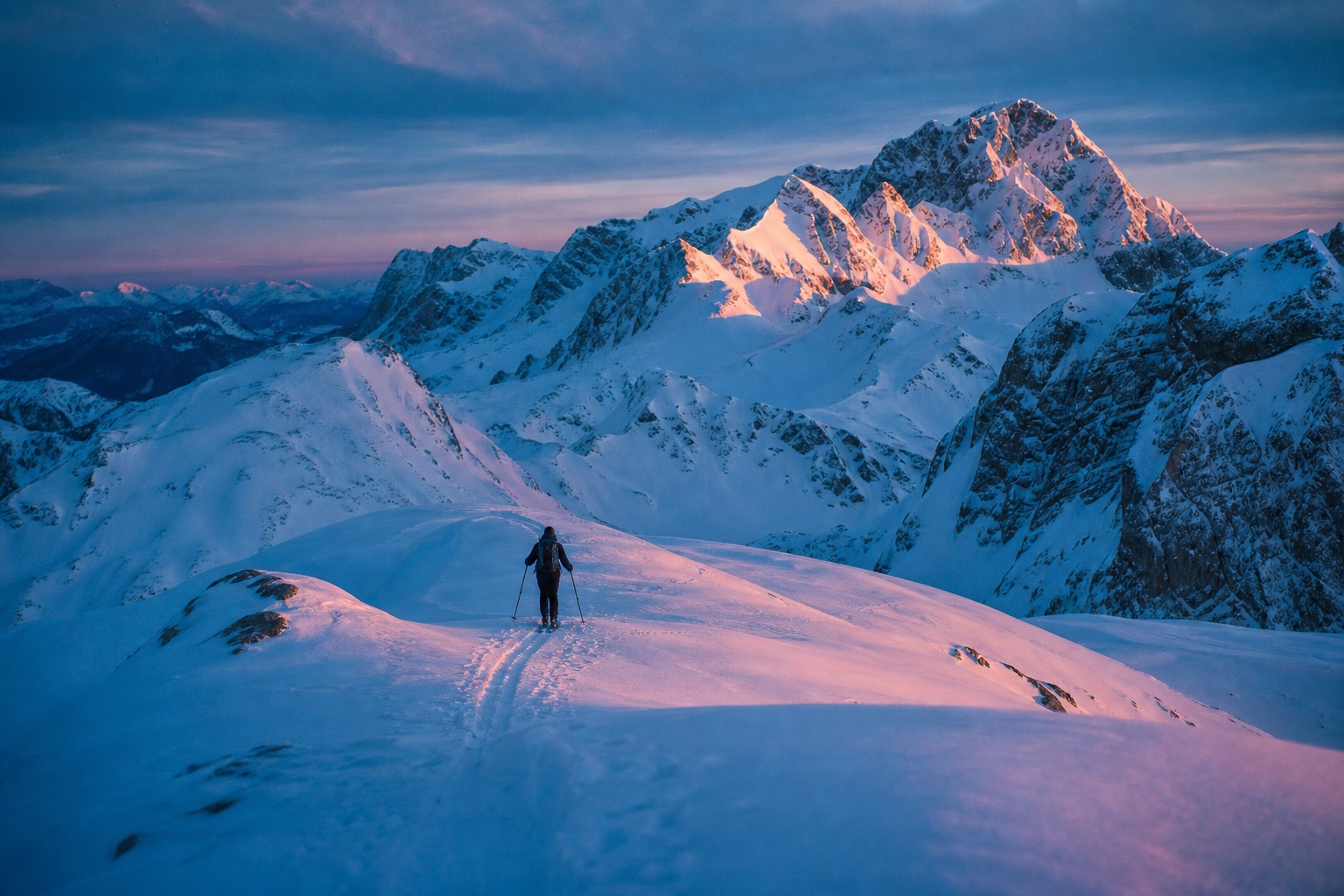 Ski touring across a pristine snow ridge in the Austrian Alps at blue hour, peaks glowing pink.