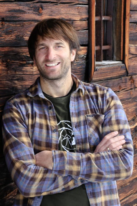 Christian, arms crossed and smiling, against a weathered alpine cabin wall.